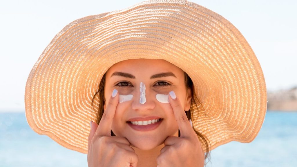 smiling woman applying sunscreen on face with wide hat on